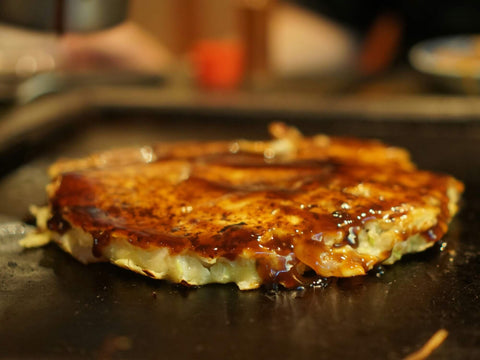 A cooked okonomiyaki sits on a grill covered in sauce and waiting for toppings