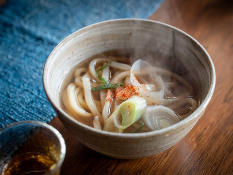 A steamy bowl of udon with green onion sits on a table next to some tea
