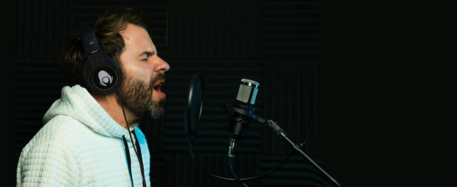 A bearded man wearing black over-ear headphones and a white hoodie sings passionately into a professional studio microphone in a soundproofed recording studio.