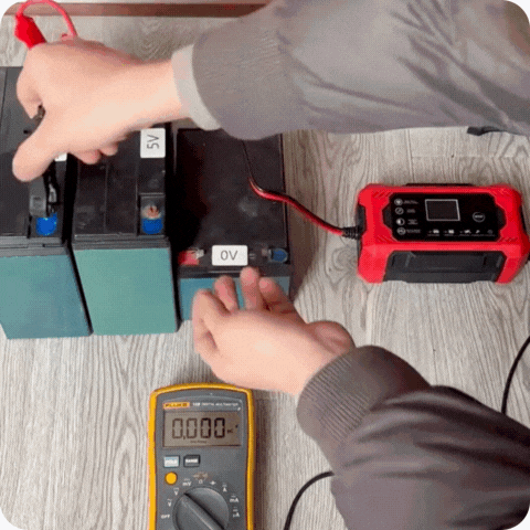 Een zwarte ndotohuis 12v universele batterijlader (6a) met rode en zwarte kabels bevestigd, verbonden met een autobatterij onder de motorkap van een auto. (Dit beschrijft het basisuitgifte en de functionaliteit)