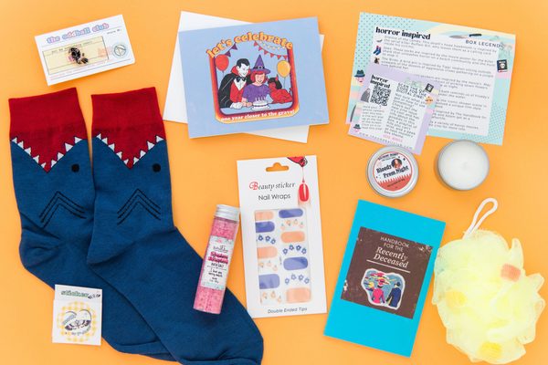 A pair of shark socks, a sarcastic birthday greeting card with a joke about death, set of nail wrap flower stickers, yellow loofa, blue notebook with handbook of the recently deceased on it, crow enamel pin, carrie bacon scented candle, and raspberry bath salt on an orange background.  