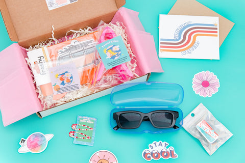 An open box with sunglasses, protective case, hair clips, and beach accessory, reflecting the chic and trendy vibe of summer.