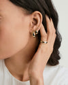 Woman wearing the Thin Dôme Ring, a simple polished gold band, gently touches her ear adorned with three small gold earrings.