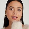 Woman with long black hair wearing chunky large gold hoop earrings and a white sweater.