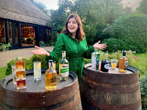 A picture of Becky Paskin in front of two barrel that have various bottles of alcohol on top