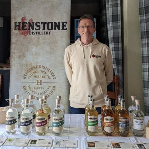 A picture of one of the owners of Henstone in front of the table with every bottle of henstone alcohol