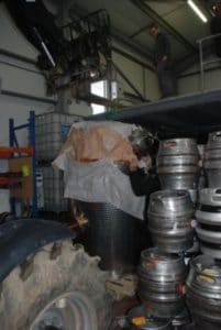 An image of an alcohol still being lifted by a small crane next to a stack of silver beer kegs