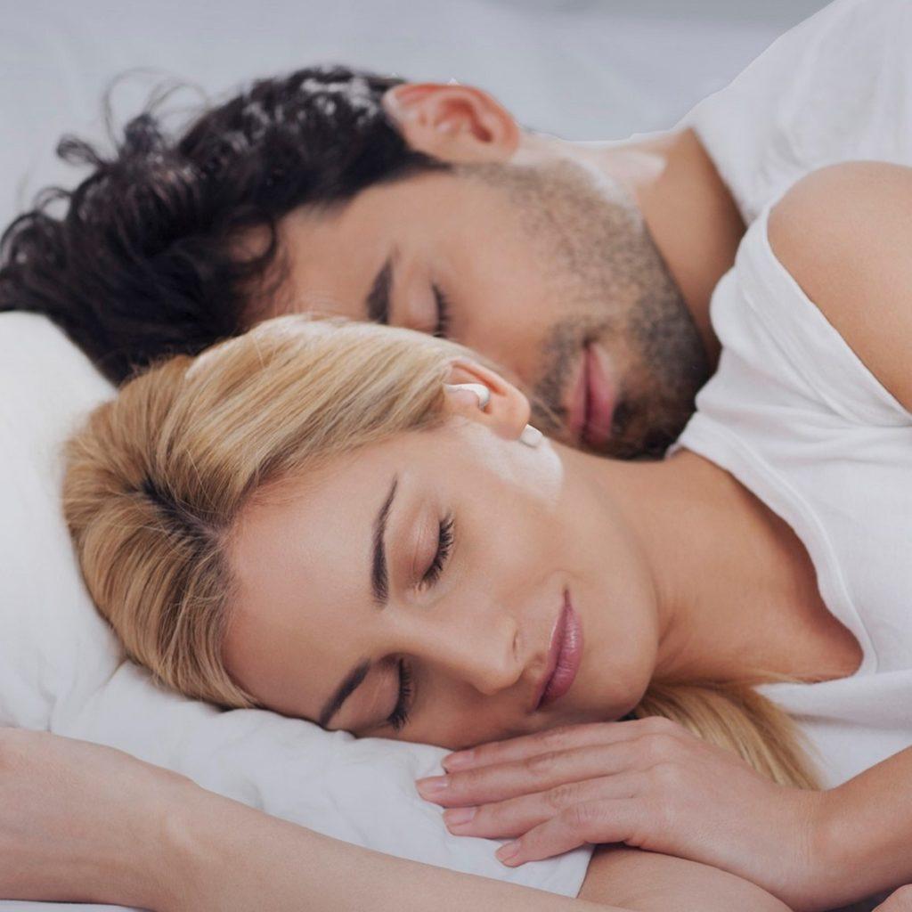 A couple snuggled together in bed sleeping peacefully. The woman is wearing noise-blocking earbuds.