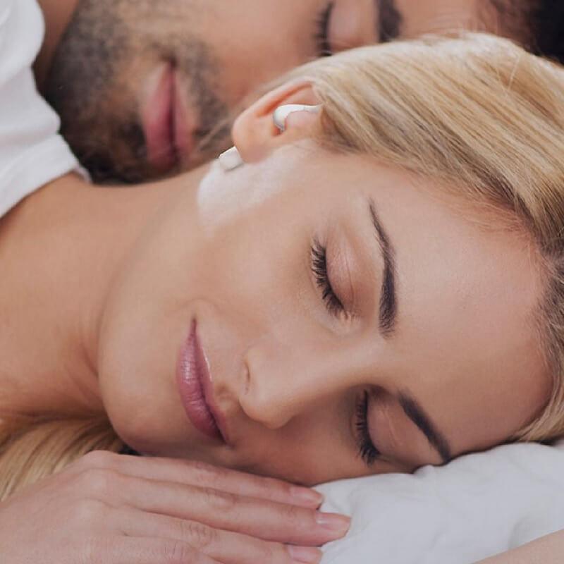A woman wearing sleep earbuds peacefully sleeps next to her snoring partner.