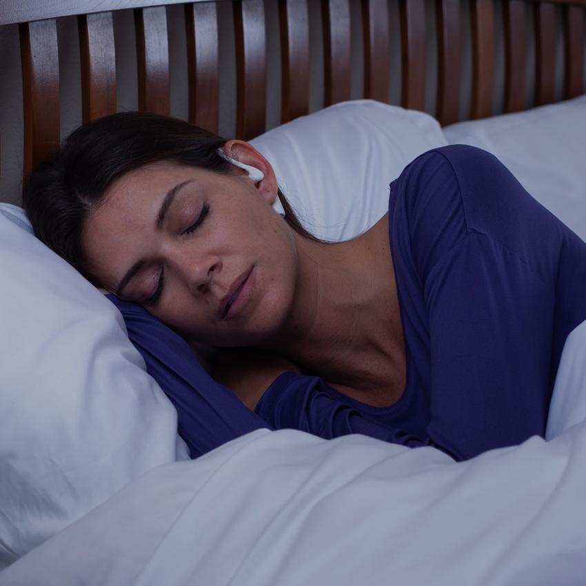 A young brunette woman wears her SoundOff Sleep Noise Masking Earbuds while sleeping soundly and calmly and deeply in bed under a duvet.