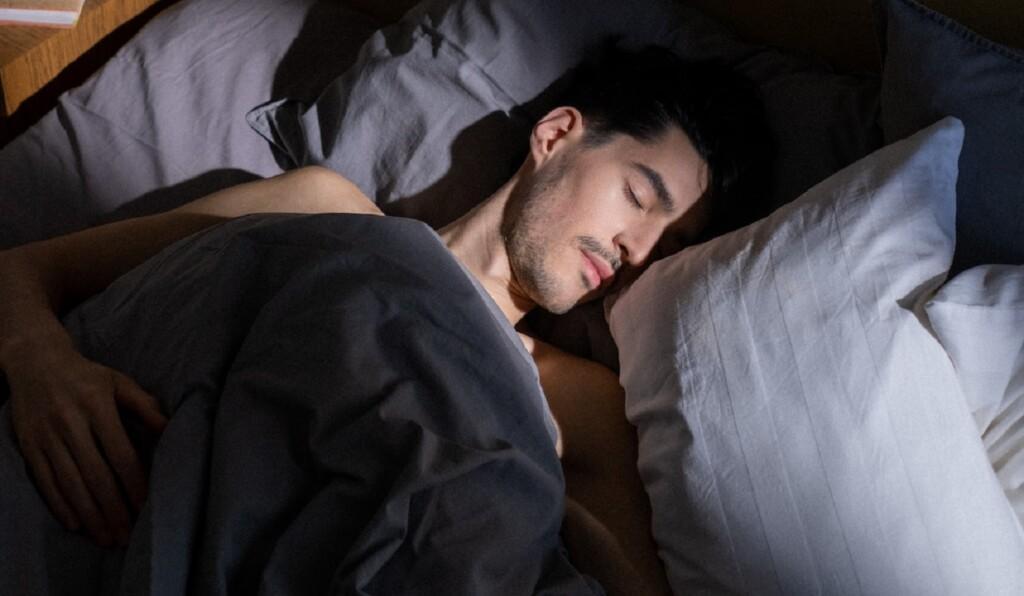 A dark-haired man sleeps deeply under a blanket. Deep sleep allows the body to repair and restore itself.