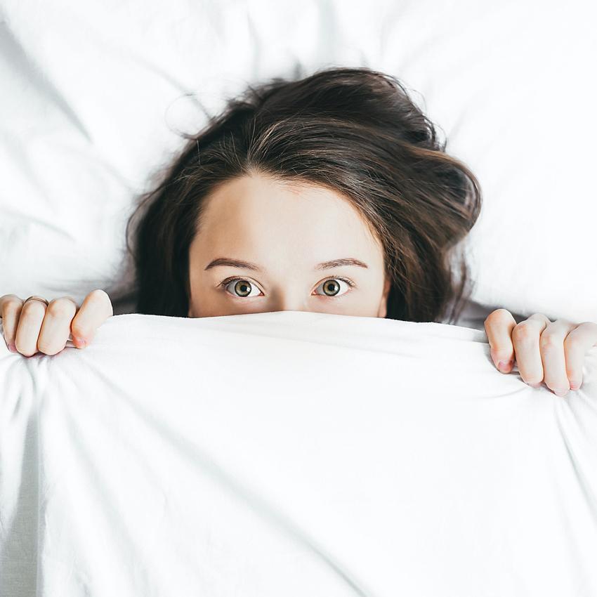 A young woman in bed stares anxiously over the top of a white comforter