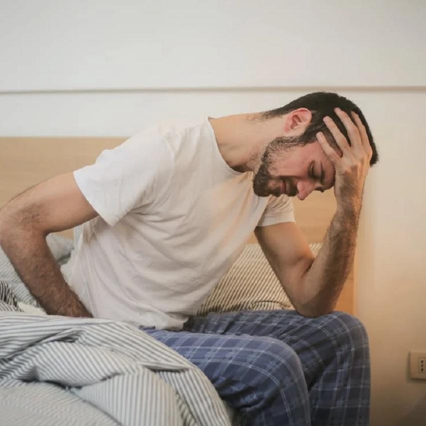 A man grimacing in distress because he can't sleep, sits on the edge of the bed, with his head in his hand