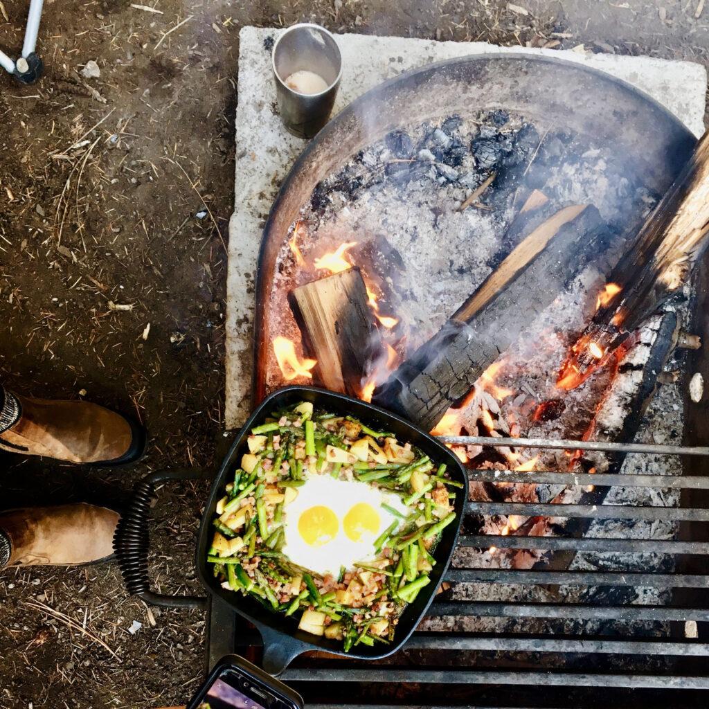 A stir fry of vegetables topped by two fried eggs cooks over a campfire. Eggs aid sleep by supplying melatonin, tryptophan, and vitamin D.