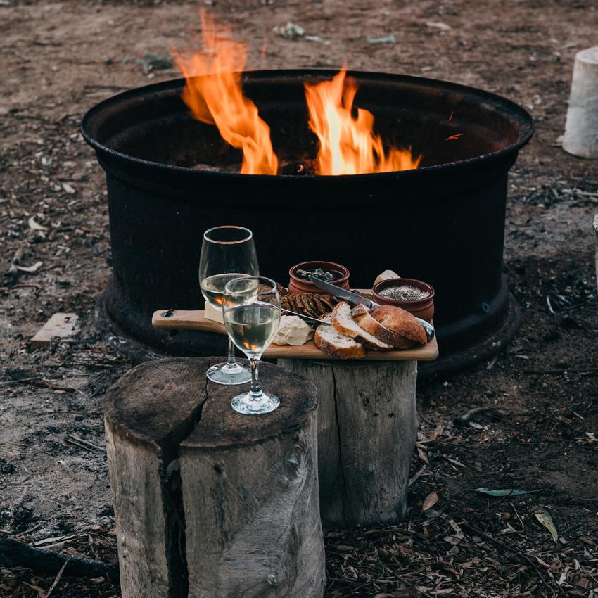 2 tree stumps in front of a small campfire hold wine glasses, and a cutting board with bread and condiments. Freeing up time for relaxing moments is good for your sleep.