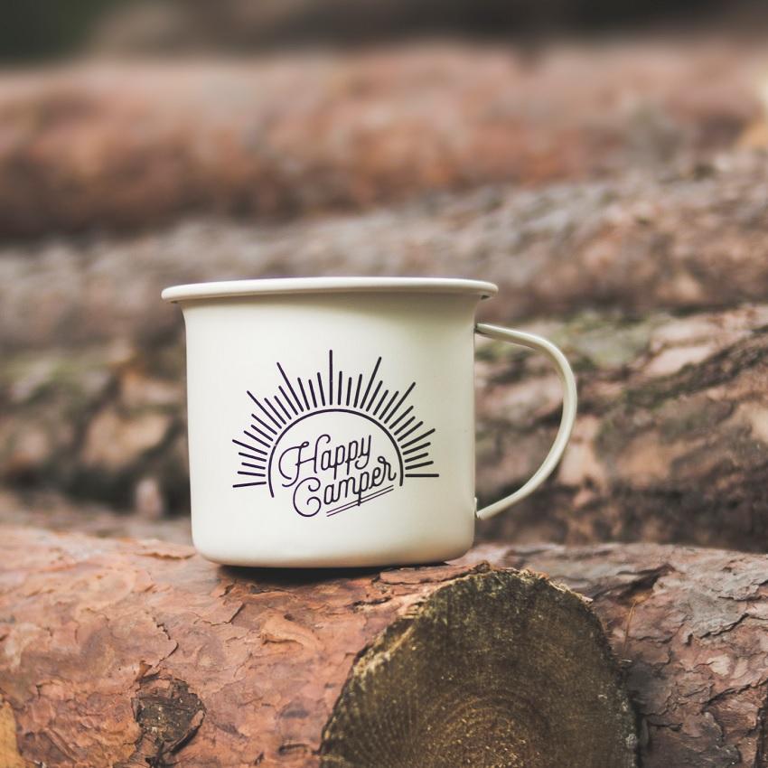 A cream-colored tin mug with the words Happy Camper rests on a stack of chopped wood. 