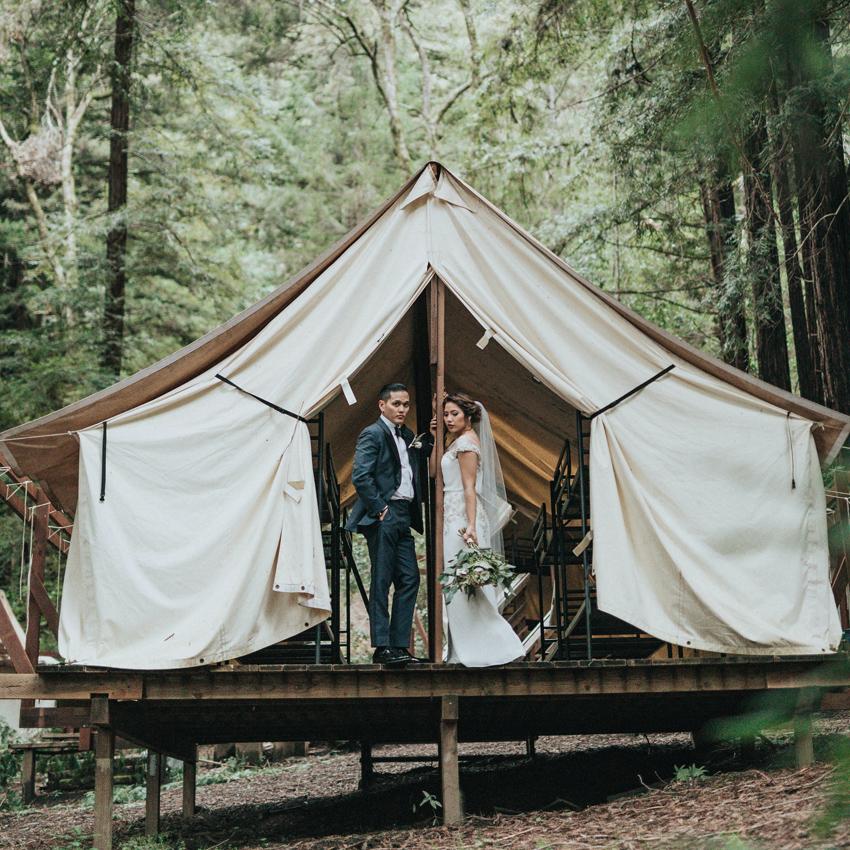 A young couple in wedding attire stands in the doorway of their glamping tent, posing for photos.