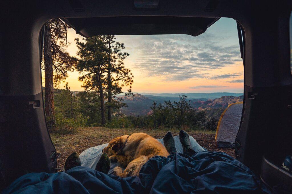 A serene mountain vista viewed from the back of a camper van. In the foreground we see two sets of feet poking out from a blanket, and a sleeping dog.