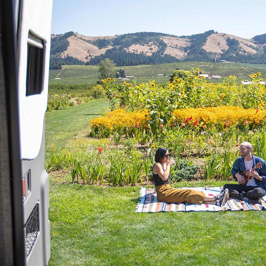 A couple relaxes outside their RV on a picnic blanket in a field with sunflowers, 