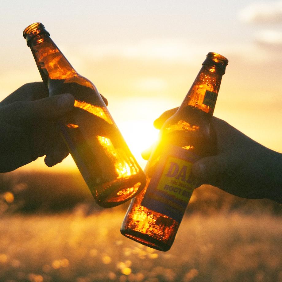 Two hands clink root beer bottles as the sun sets. over a grassy meadow. Avoiding alcohol at bedtime improves sleep quality.