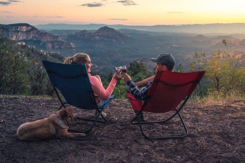 A young couple in camp chairs clink wine glasses as they enjoy the sunset and time alone with each other and their dog.