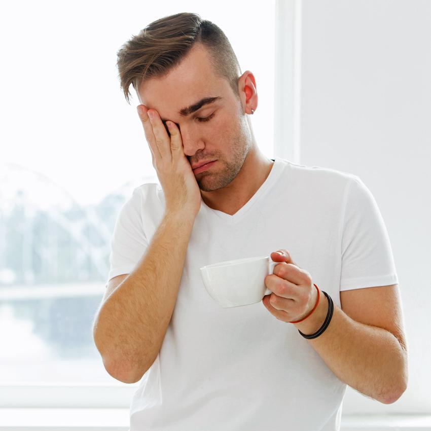 A sleepy young man in white tee-shirt holds a white coffee mug and rubs his face. A 20-minute "coffee nap" can help keep you alert longer than a nap alone.