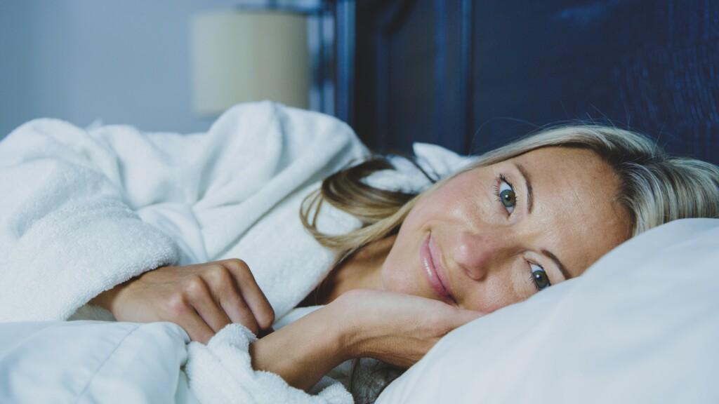 A smiling blond woman lies in a white bed, looking refreshed and happy. 