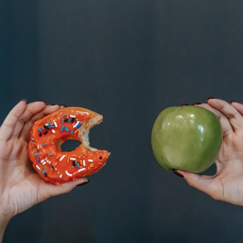 Two hands.  One holding a donut and the other hand holding a green apple