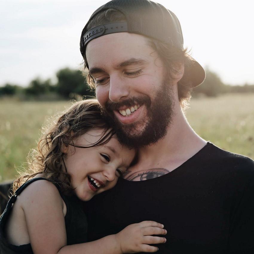 An off-grid father holds his daughter as the two smile and laugh while walking in a grassy field.