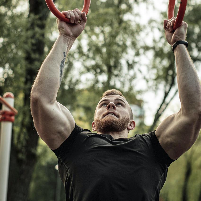 A young ginger-haired man with bulging muscles does pull ups on rings as resistance training. 