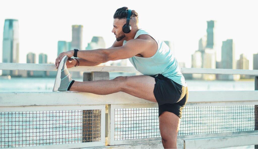 A young man wearing headphones pauses his run across a bridge to stretch and flex his legs. He looks out over the water to a cityscape.