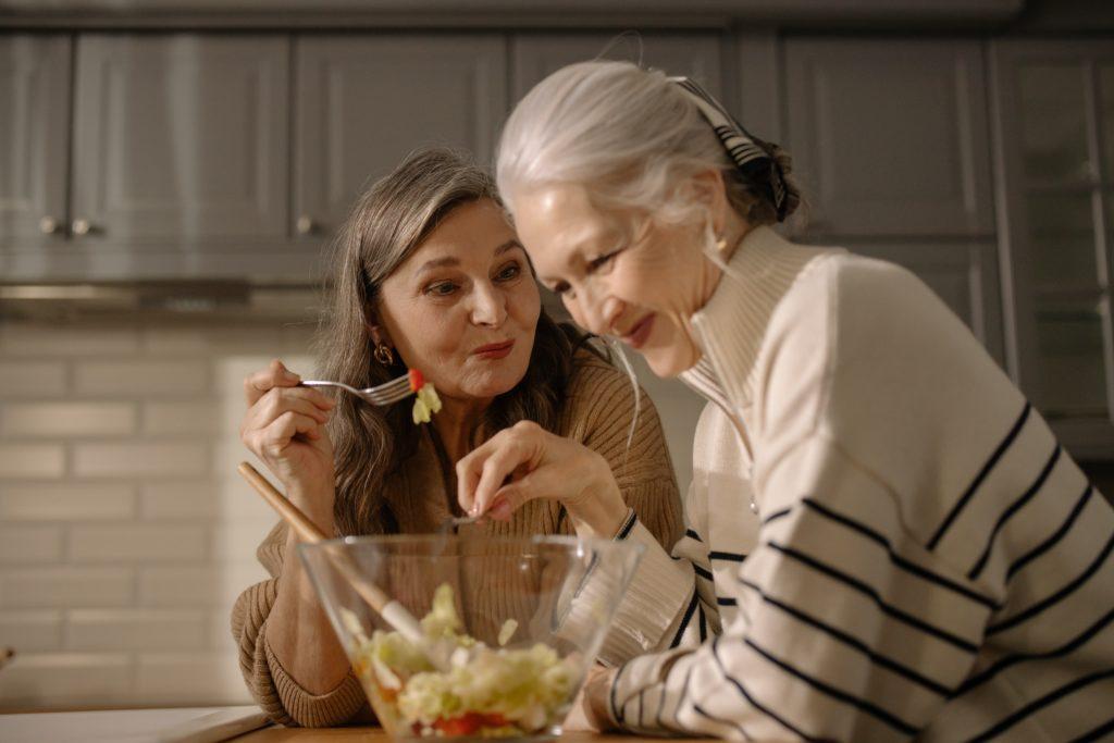 Two older ladies at kitchen counter eating salad and laughing