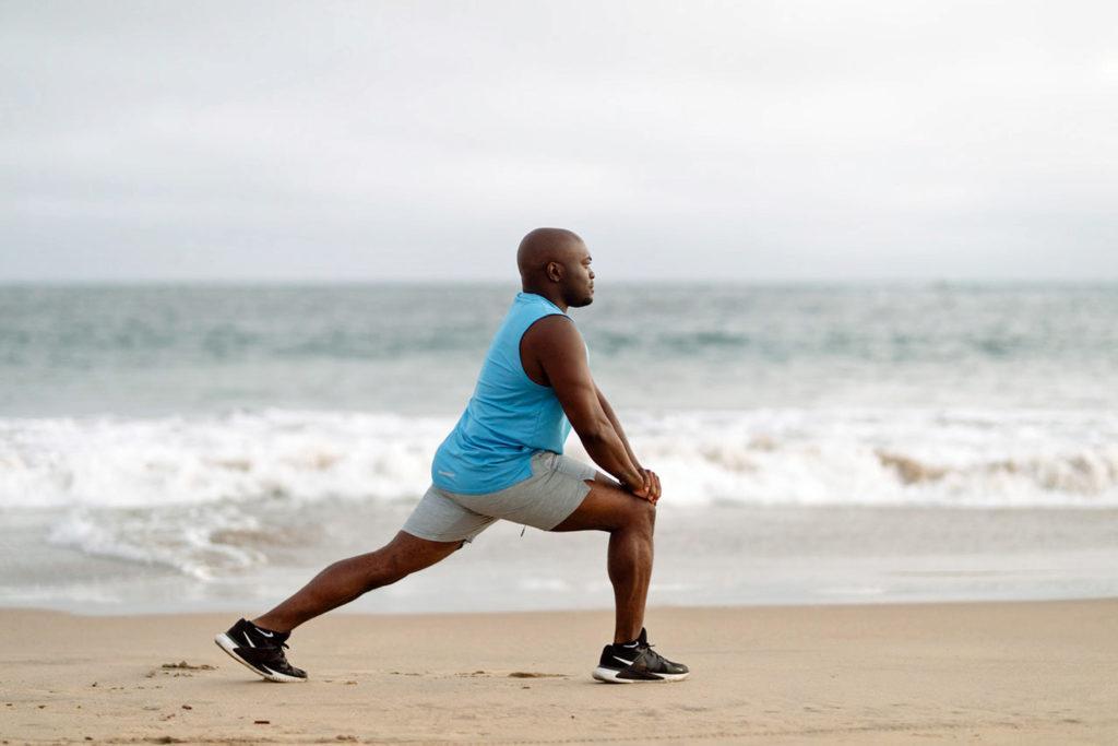 Man in blue shorts doing leg stretch on the sand with ocean in background