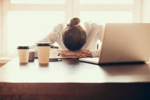 A woman face-down on her desk with laptop and multiple cups of coffee