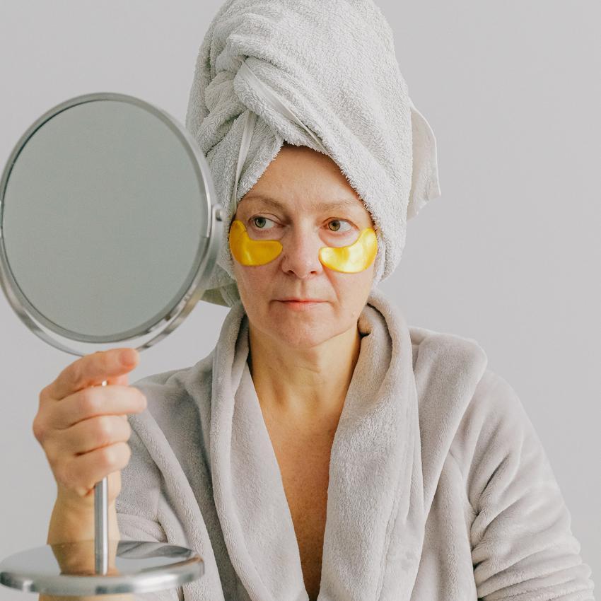 Woman in bathrobe and towell on hair looking in hand held mirror. Woman has eye mask under her eyes to help with puffy eyes