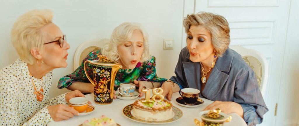 3 older ladies sitting at table with tea cups and blowing out candles on a cake