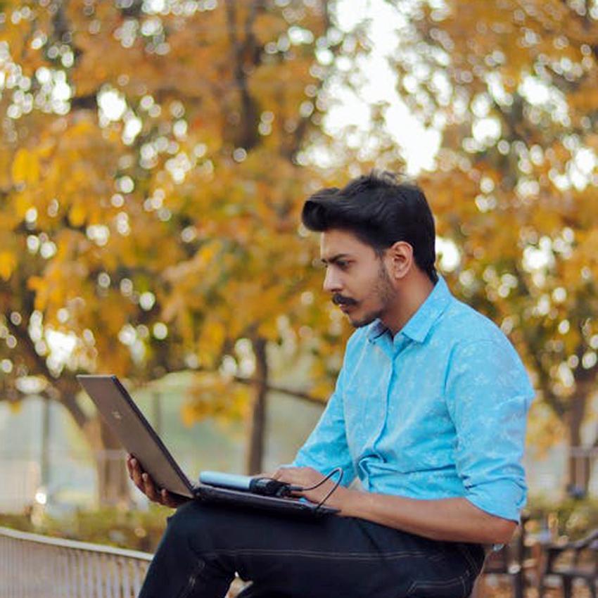 A dark haired man works outside in the park on his laptop