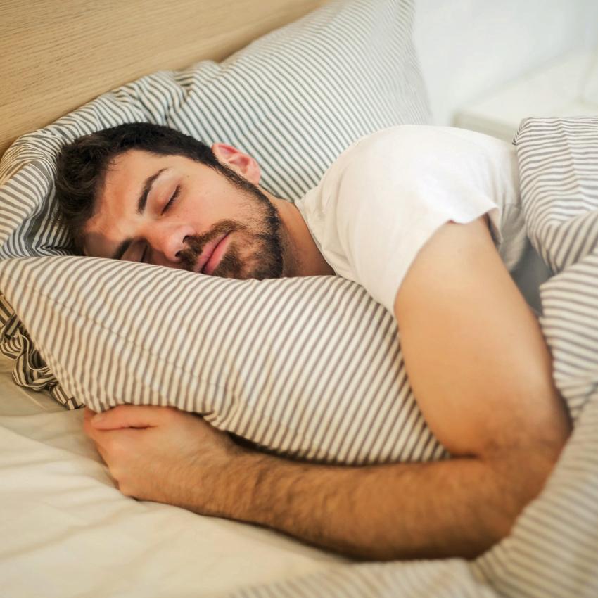 A young man sleeping with his arm around a pillow as morning light starts creeping into his bedroom..