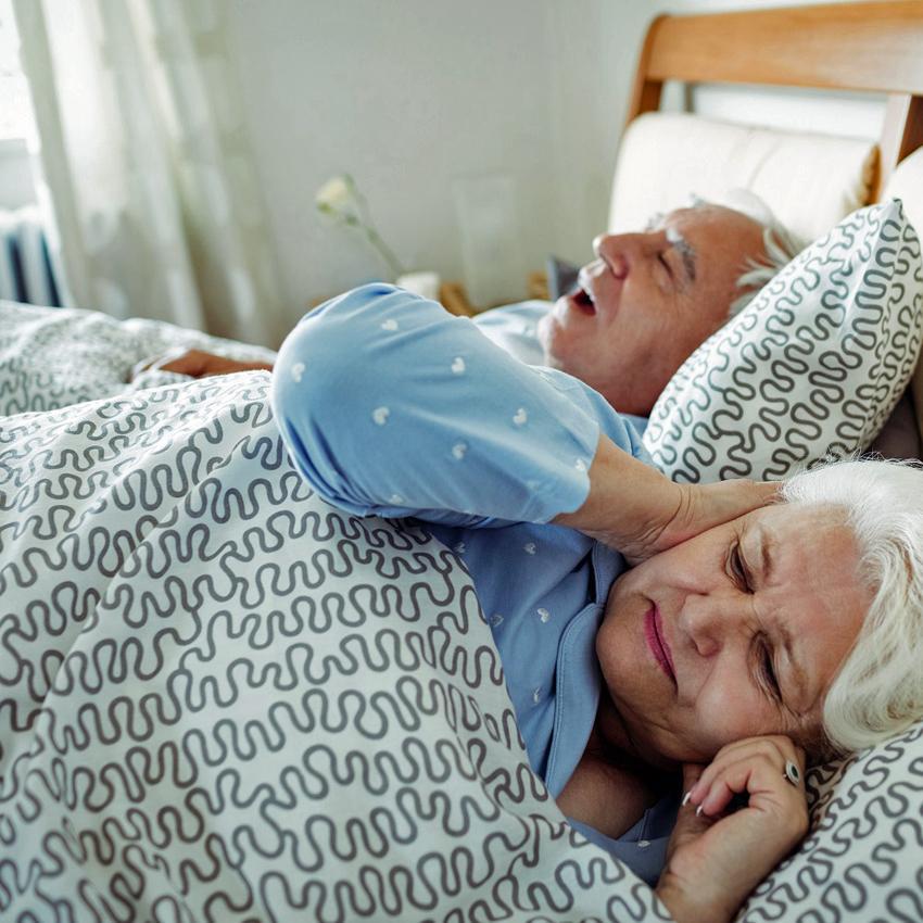 Elderly woman in bed with husband is covering her ears to block out his snoring