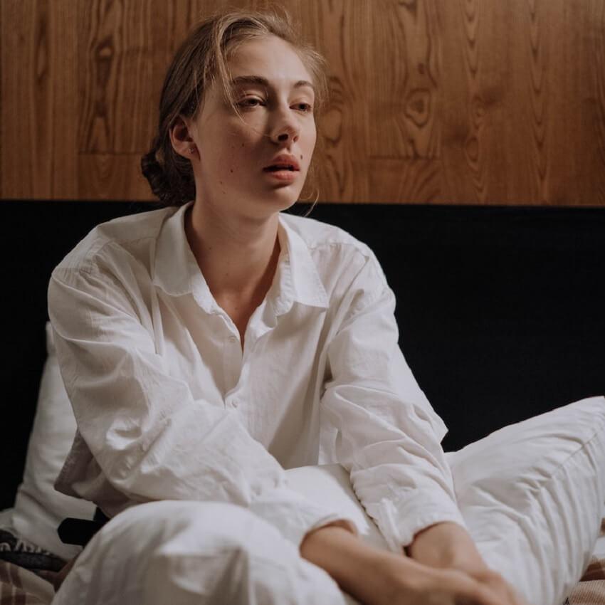 An exhausted blond woman sits on bed, looking dazed and not ready to start her day.