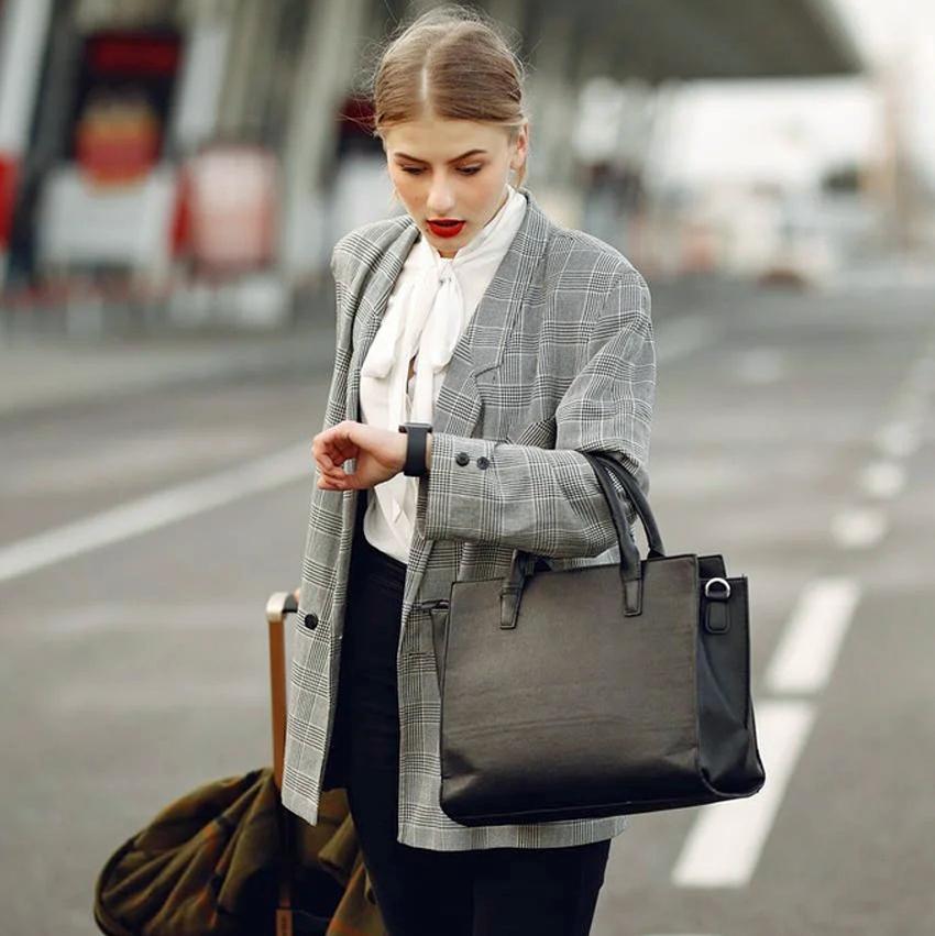 A woman traveling in business suit crosses street with luggage, looking at watch with worried expression as if running late