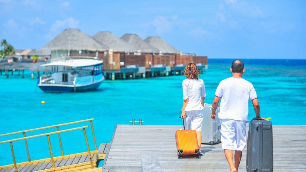A man and woman traveling, walk along a dock, pulling their suitcases as they head toward their vacation huts.