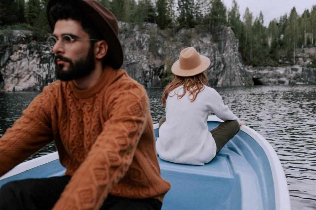 A couple in a small boat on a river sit facing away from each other. Man stares off into the distance