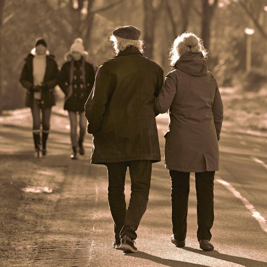 An elderly couple strolls arm-in-arm along a tree-lined path, approaching other people who are also out walking. 