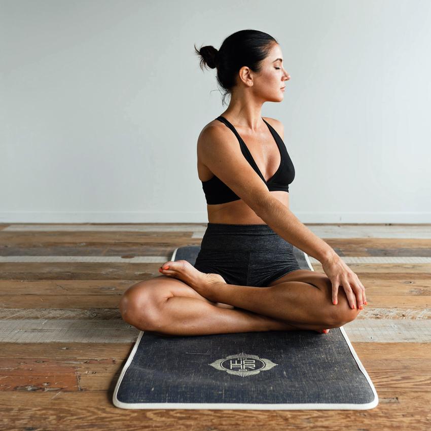 A cross-legged yoga stretch is performed by a young woman on a yoga mat.