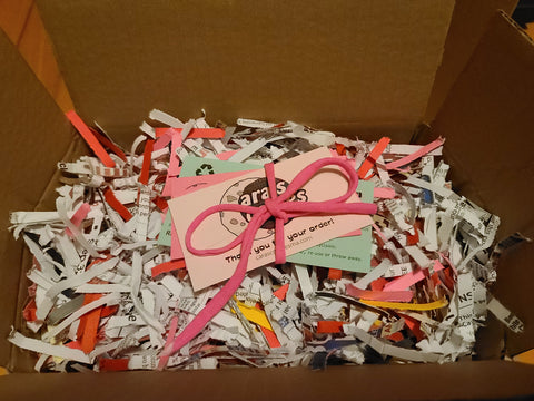 open box with multicolored shredded paper filling, colored paper cards, and a pink ribbon bow