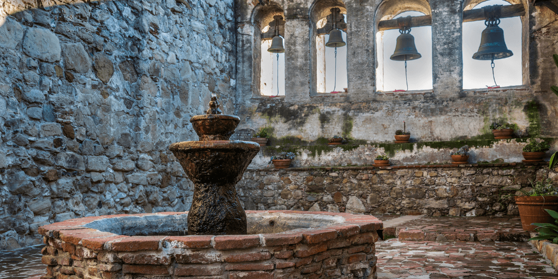 San Juan Capistrano Mission Bells and Fountain: A serene courtyard scene featuring the mission's iconic bells, framed by vibrant gardens and a tranquil fountain, showcasing the essence of California's rich history.