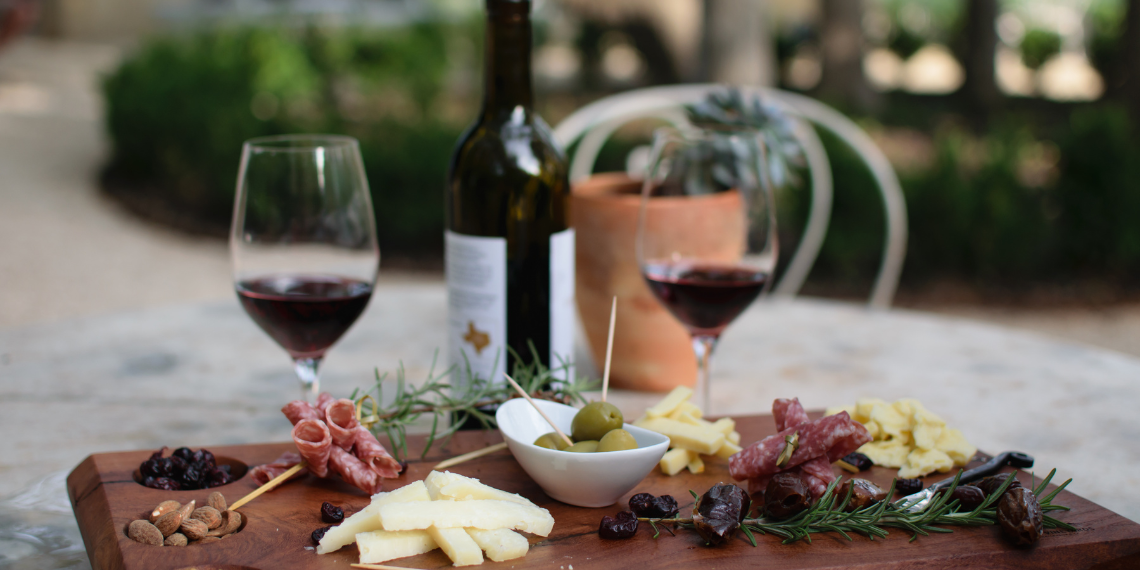 An alfresco wine dinner in San Juan Capistrano featuring a beautifully arranged charcuterie board, adorned with an assortment of artisanal cheeses, cured meats, and savory accompaniments, complemented by glasses of fine wine, set against the backdrop of a serene outdoor setting.