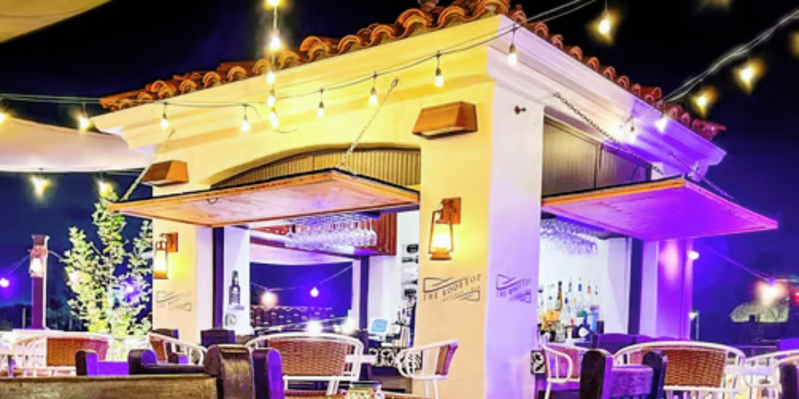 Glowing lights illuminate The Rooftop Kitchen + Bar, casting a warm and inviting ambiance over the vibrant rooftop setting, perfect for socializing and enjoying drinks and cuisine under the night sky.