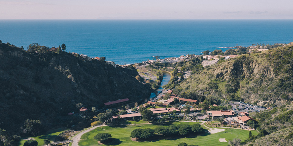 Aerial view of The Ranch at Laguna Beach property, showcasing a serene creek winding through a picturesque golf course with a stunning backdrop of the ocean on a sunny day, creating a harmonious blend of nature and luxury.
